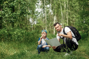Fototapeta premium A man with a backpack, a father and his son on a hike look at the map, walking during walks in the woods. Family life, pastime with the family, the upbringing of children.