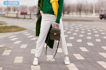 Street, bright style. A young girl in white trousers, warm greens polto. White Sneakers. Yellow scarf. Green jacket. A brown handbag. Details.