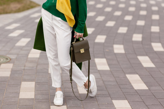 Street, bright style. A young girl in white trousers, warm greens polto. White Sneakers. Yellow scarf. Green jacket. A brown handbag. Details.