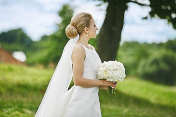 Beautiful and elegant young blonde woman in stylish wedding dress with bouquet of white flowers in her hands posing outdoors