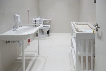 A bathroom with a toilet bowl for the disabled, a sink and a changing table for babies
