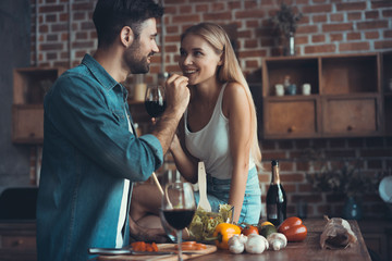 Beautiful young couple is feeding each other and smiling while cooking in kitchen at home.