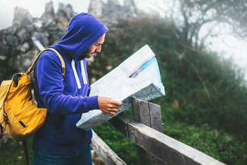 Hipster young man with backpack holding in hands and looking on map spain of foggy mountain,...