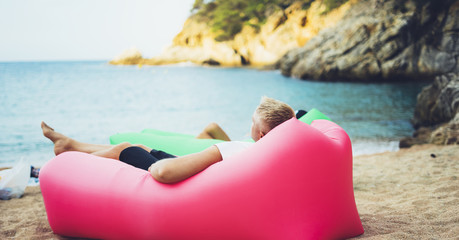 young hipster relaxing on coastline beach on inflatable lazy air pouffe sofa, person tourist enjoy sunny day on background coast sea waves, seascape perspective vacation, summer seashore