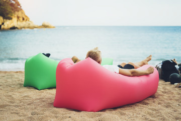 young hipster relaxing on coastline beach on inflatable lazy air pouffe sofa, person tourist enjoy sunny day on background coast sea waves, seascape perspective vacation, summer seashore
