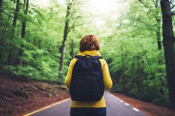 tourist traveler with backpack into road at summer green forest, view back girl hiker in yellow hoody looking and enjoying the breath of fresh air in trip, relax holiday concept, blurred background - obrazy, fototapety, plakaty