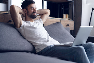 Typing new blog post. Side view of handsome young man using his laptop with smile while sitting on the couch at home.
