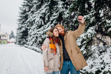Young couple in love make joint photo of themselves on mobile phone. The guy holds smartphone on his elongated hand and makes selfie photo on background of park with coniferous trees in winter