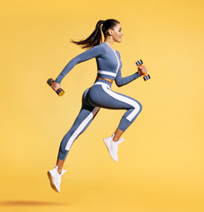 Sporty woman jumping with dumbbells. Photo of active woman in sportswear on yellow background. Dynamic movement. Side view. Sport and healthy lifestyle - obrazy, fototapety, plakaty
