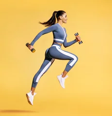 Badezimmer Foto Rückwand Sporty woman jumping with dumbbells. Photo of active woman in sportswear on yellow background. Dynamic movement. Side view. Sport and healthy lifestyle © Romario Ien
