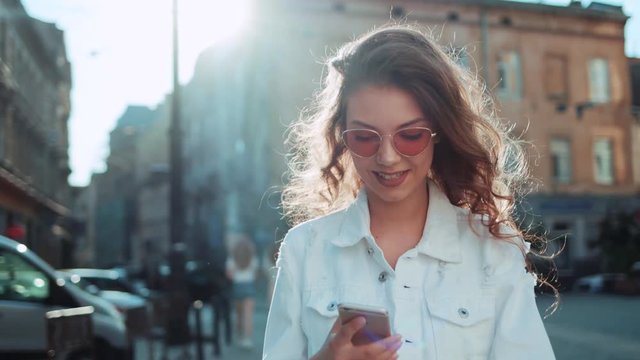 Smiling curly woman wearing trendy sunglasses walks down the central city street and uses her phone. Pretty summer woman in white jacket walks down the street looking at her mobile phone