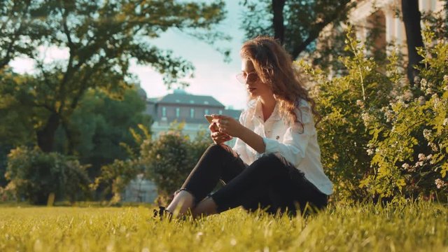 Beautiful curly female student with modern sunglasses has a break, sits on the grass in the park near the university, actively uses her smartphone. Relaxation, browsing the internet, being online