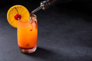 Making Tequila Sunrise Cocktail. Grenadine slowly poured into a glass of ice, orange juice and...