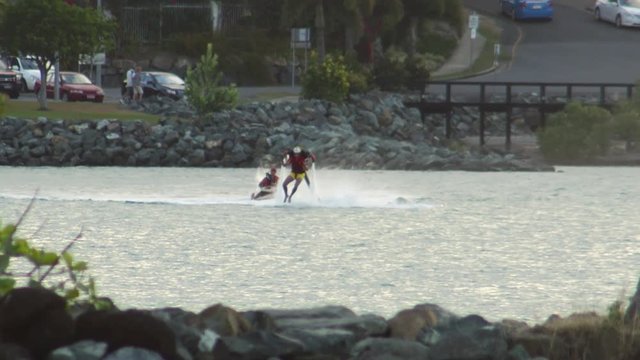 A wide shot of a man on a water jet pack as he moves forward on the air. A jet ski follows him.