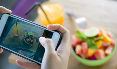 close up female hand with mobile phone screen taking picture of fruit salad and orange juice for sharing on internet social media app in healthy nutrition