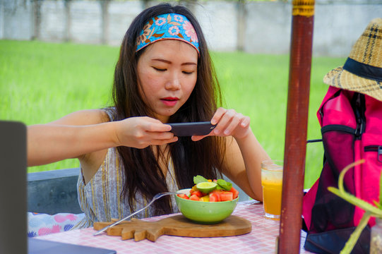 young happy and pretty digital nomad Asian Chinese woman taking picture of fruit salad with mobile phone networking on internet social media