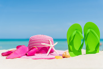 Fototapeta na wymiar Beach accessories including flip flop, starfish, beach hat and sea shell on sandy beach, green sea and blue sky background for summer holiday and vacation concept.