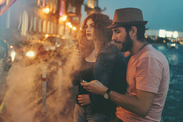 Loving couple of hipsters on date. Guy in hat and T-shirt is hugging his girlfriend. Girl with curly red hair smokes electronic cigarette. They are in streets of evening city.