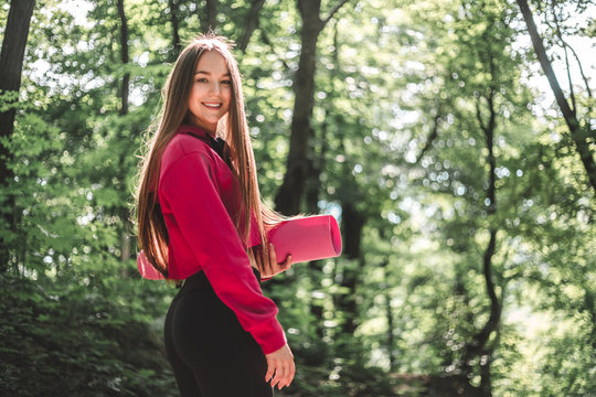 Sporty woman in hoodie with a fitness mat on training outdoors. Cropped image of a young athletic woman with in a sporty sweatshirt with a hood holding a sports mat on a forest path.