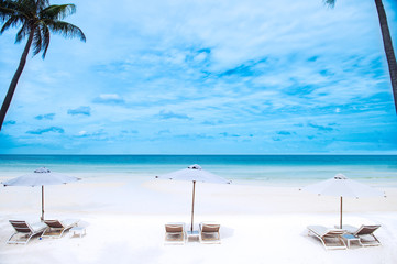 An empty white sand beach with three empty sun beds and parasols and an empty ocean with blue sky