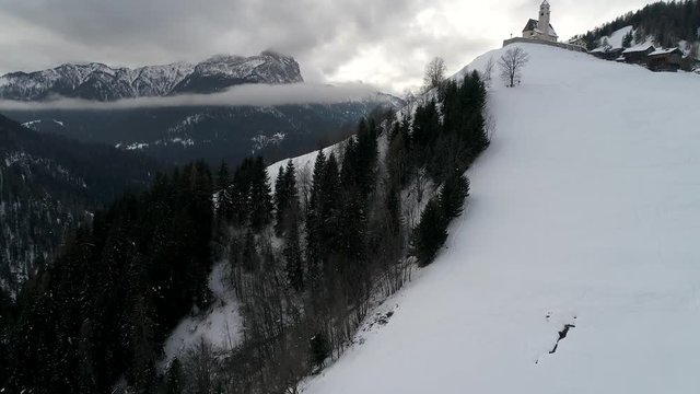 aerial drone flying above small church in italian alps at cludy winter day