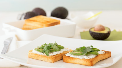 Roasted toasts with cream cheese and slices of avocado, leafs of rucola and pink Himalayan salt. Front view, low angle, focus on the centre of both breads. White wood background.