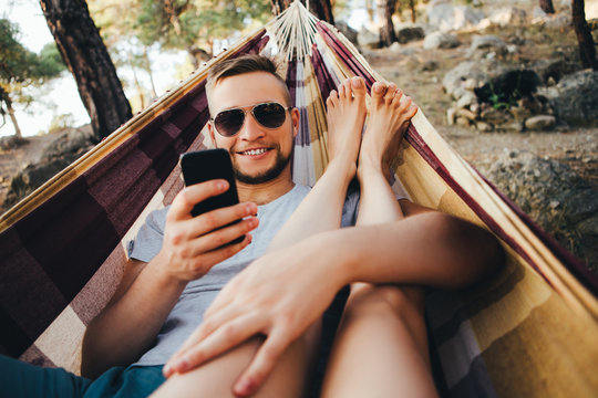 Time of laziness. A man is lying in a hammock and taking pictures of his girlfriend on the phone.
