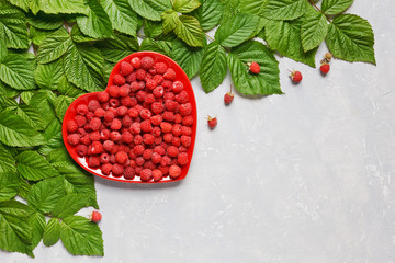 Fresh juicy raspberries on red heart shaped plate and pattern of raspberry leaves. Top view, close-up on  light gray background