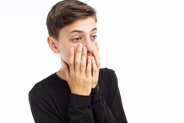 Photo emotional teenager, guy in black t-shirt, shows the emotions of fear, closing his mouth with his hands, on a white background, for advertising text insertion