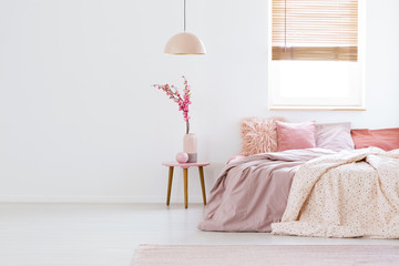 Lamp above table with flowers in pink pastel bedroom interior with window above bed. Real photo - obrazy, fototapety, plakaty