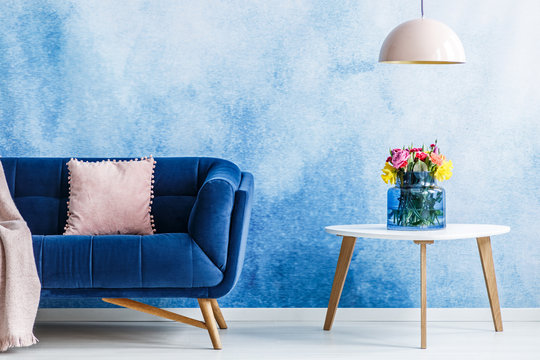Comfortable plush settee with pastel cushion and a side table with fresh flowers in a vase against blue and white ombre wall in a living room interior. Lamp hanging over the table. Real photo.
