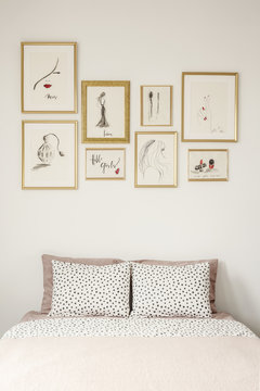 Stylish drawings gallery in golden frames above a cozy double bed with polka dot sheets in a white apartment room interior of a beauty lover