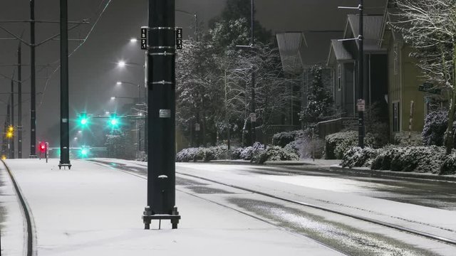 Traffic moves on wet streets with light snow and light rail power poles in foreground at night. Light rail trains pass by.