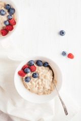 A bowl of oatmeal porridge with blueberries and raspberries on white wooden table. Top view.