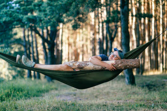 Odd weird strange unusual male person. Outlandish funny crazy foolish man sleeping in hammock with huge wooden log at nature among trees.  Wood and forest lover. With beam in bed.  Leisure lifestyle.