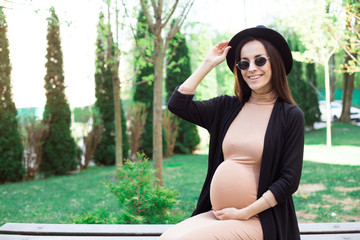 Stylish Pregnant in Hat and Sunglasses Sitting on a Bench in the City Park and Relax