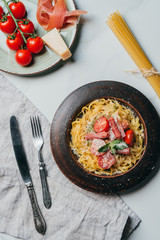 top view of plates with pasta and jamon, parmesan and jamon on marble table with kitchen towel, fork, knife and raw spaghetti