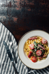 elevated view of pasta with mint leaves, jamon and cherry tomatoes covered by grated parmesan on plate at table with kitchen towel, knife and fork