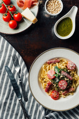 top view of pasta with mint leaves, jamon and cherry tomatoes covered by grated parmesan on plate at table with kitchen towel, knife, fork, pine nuts and pesto sauce in bowl