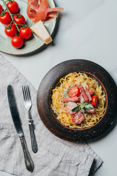 elevated view of plates with pasta and jamon, parmesan and cherry tomtatoes on marble table with kitchen towel, fork and knife