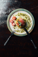 elevated view of pasta with mint leaves, jamon, pine nuts, pesto and cherry tomatoes covered by grated parmesan on plate with fork and knife at table
