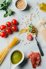 elevated view of fork wrapped by pasta surrounded by pine nuts, pesto, parmesan, raw spaghetti, cherry tomatoes, salt, olive oil and jamon on marble table