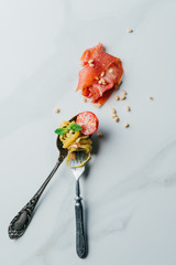 elevated view of spoon and fork wrapped by pasta with mint leaves, pine nuts, jamon and cherry tomatoes covered by grated parmesan on marble table