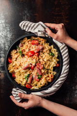 cropped image of woman holding pasta with jamon, cherry tomatoes, mint leaves covered by grated parmesan in pan wrapped by kitchen towel over table