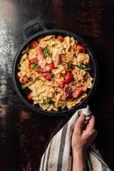 partial view of woman holding pasta with jamon, cherry tomatoes, mint leaves covered by grated parmesan in pan wrapped by kitchen towel over table
