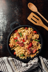 elevated view of pasta with jamon, cherry tomatoes, mint leaves covered by grated parmesan in pan on table with kitchen towel, wooden spoon and spatula