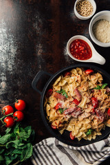 elevated view of pasta with jamon, pine nuts, sauce, cherry tomatoes, mint leaves covered by grated parmesan in pan surrounded by ingredients and kitchen towel on table