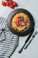 top view of pasta with mint leaves, jamon and cherry tomatoes covered by parmesan on plate at marble table with kitchen towel, knife and fork