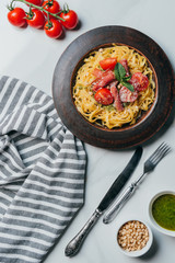 view from above of pasta with mint leaves, jamon and cherry tomatoes covered by parmesan on plate at marble table with kitchen towel, knife, fork, pine nuts and pesto sauce in bowl