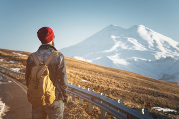Bearded tourist hipster man in sunglasses with a backpack stand back on a roadside bump and watching the sunset against the background of a snow capped mountain